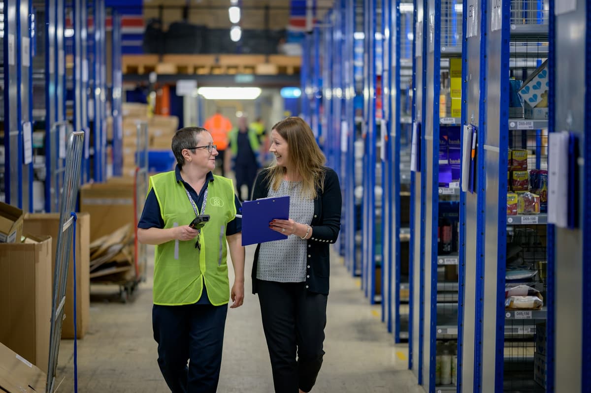 Two colleagues talking as they walk through a warehouse aisle with a clipboard